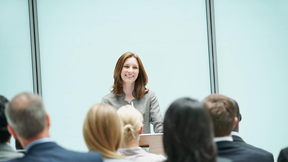 A woman presenting to a room full of seated people in business attire